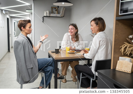 Businesswomen communicating, smiling, and standing together at kitchen counter during company lunch break. Communicating, lunch break, kitchen, teamwork, office, businesswomen. Businesswomen communicating, smiling, and standing together at kitchen counter during company lunch break. Communicating, lunch break, kitchen, teamwork, office, businesswomen. 138345505
