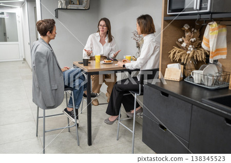 Two women colleagues eating together at counter in work kitchen during lunch break at office. Eating, kitchen, lunch break, colleagues, teamwork, office. Two women colleagues eating together at counter in work kitchen during lunch break at office. Eating, kitchen, lunch break, colleagues, teamwork, office. 138345523