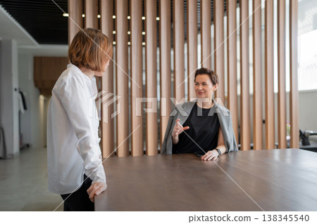 Women having collaborative conversation at wood table in bright contemporary business office. Conversation, collaboration, wood table, business, office, women. Women having collaborative conversation at wood table in bright contemporary business office. Conversation, collaboration, wood table, business, office, women. 138345540