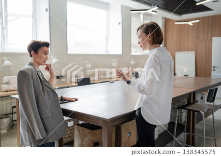 Women discussing at office desk, one seated and one standing in bright workspace with wood accents. Discussion, desk, teamwork, office, standing, women. 138345575
