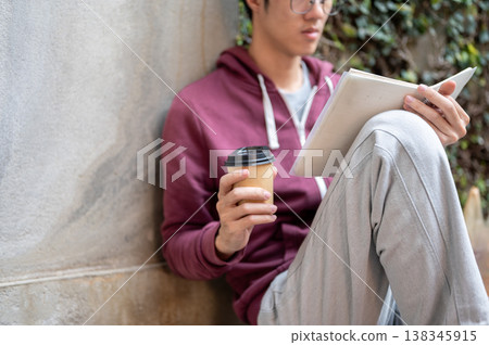 Glasses young asian man holding coffee reading a book while sitting knee up leaning on concrete wall 138345915