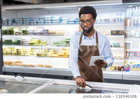 African american man wearing an apron and glasses standing in a supermarket aisle, holding a tablet computer while checking inventory in a frozen food freezer, working efficiently to manage stock African american man wearing an apron and glasses standing in a supermarket aisle, holding a tablet computer while checking inventory in a frozen food freezer, working efficiently to manage stock 138346106