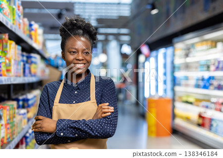 African american woman with an apron and folded arms standing confidently in a supermarket aisle, representing a dedicated worker in the grocery business 138346184