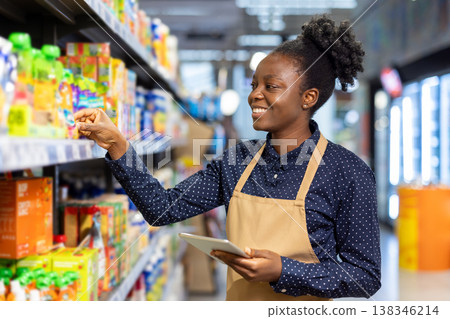 African american woman employee in apron maintaining product display on supermarket shelves, checking inventory with a digital tablet while working in a grocery store African american woman employee in apron maintaining product display on supermarket shelves, checking inventory with a digital tablet while working in a grocery store 138346214