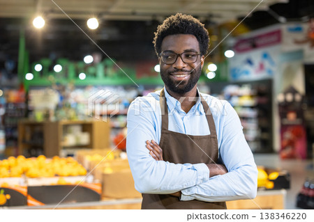 Young african american grocery store owner wearing an apron, arms crossed and smiling confidently at the camera while managing his small business among fresh produce 138346220