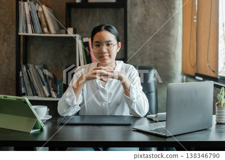 Young woman in glasses working at a desk with a laptop and tablet in a modern office setting. Young woman in glasses working at a desk with a laptop and tablet in a modern office setting. 138346790