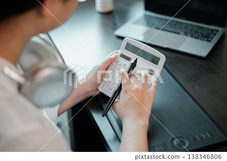 Close-up of a person using a calculator with a pen, next to a laptop and headphones on a desk. Close-up of a person using a calculator with a pen, next to a laptop and headphones on a desk. 138346806