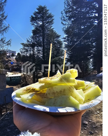 Hand holding a plate of spiced apple slices against tall pine trees and blue sky in Kufri, combining food, travel, and nature elements. Hand holding a plate of spiced apple slices against tall pine trees and blue sky in Kufri, combining food, travel, and nature elements. 138347117