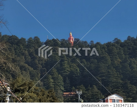 Distant view of a towering hilltop statue rising above dense pine forests in Shimla, captured under a clear blue sky, showcasing spiritual landmarks within Himalayan nature. 138347118