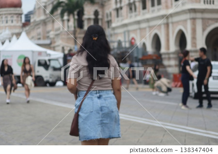 Woman walking through a public square in Kuala Lumpur, Malaysia, with colonial-era buildings and daily city life captured from a candid rear perspective. 138347404