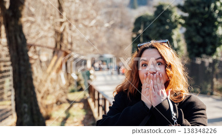 Young Slavic woman with red hair covering mouth in surprise while standing outdoors in park on sunny day expressing emotions shock and excitement lifestyle moment 138348226