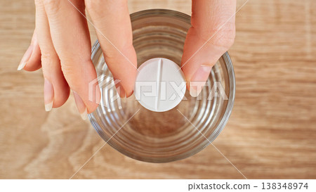 Close-up Female Hand Holding Tablet Above Glass Of Water, Top View. Medication, Supplement Dosage, Preparation And Daily Healthcare Routine Concept. Close-up Female Hand Holding Tablet Above Glass Of Water, Top View. Medication, Supplement Dosage, Preparation And Daily Healthcare Routine Concept. 138348974