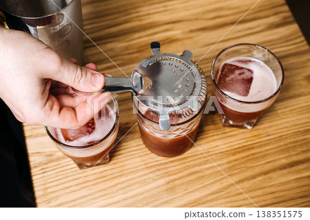 Hand holds metal strainer over glass filled with red liquid and ice alongside two other drinks. Health conscious drinking, alcohol free ritual, mindful celebration. 138351575