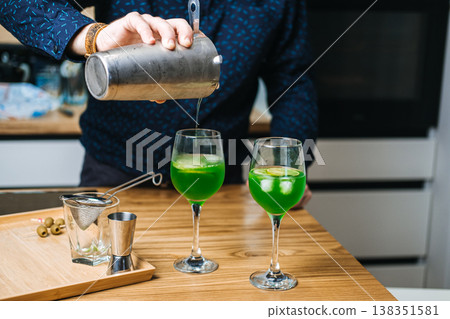 Mixing glass containing green liquid and cucumber slices sits on wooden counter next to metal shaker and lime. home bartending, craft green cocktails, mixology trend, DIY cocktail recipes Mixing glass containing green liquid and cucumber slices sits on wooden counter next to metal shaker and lime. home bartending, craft green cocktails, mixology trend, DIY cocktail recipes 138351581