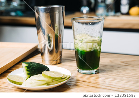 Mixing glass containing green liquid and cucumber slices sits on wooden counter next to metal shaker and lime. cucumber lime mocktail, refreshing garden drinks, citrus cucumber water Mixing glass containing green liquid and cucumber slices sits on wooden counter next to metal shaker and lime. cucumber lime mocktail, refreshing garden drinks, citrus cucumber water 138351582