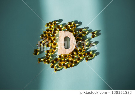 Wooden letter D rests among cluster of yellow gel capsules on blue background in sunlight. Vitamin D deficiency, proactive health, wellness routine. 138351583