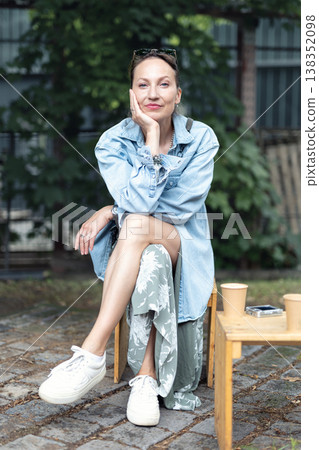 Outdoor portrait confident young adult woman sit casually urban park environment calm, elegance mindfulness modern lifestyle. Natural light greenery denim outfit relaxed pose  authentic feminine mood. 138352098