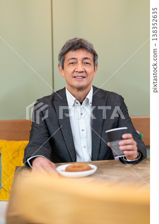 A senior businessman smiles as he takes a break at a cafe, enjoying coffee and a donut. 138352635