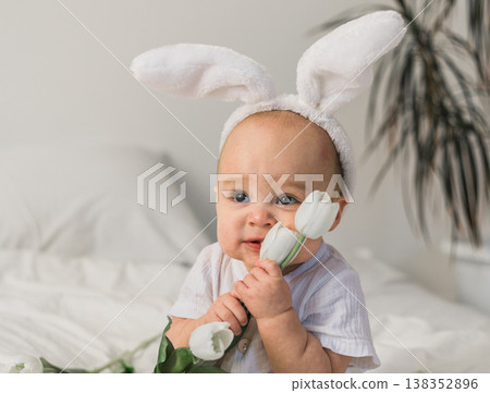 Cute baby with rabbit ears on his head and tulips at home on the bed. Easter and spring time concept. Cute baby with rabbit ears on his head and tulips at home on the bed. Easter and spring time concept. 138352896