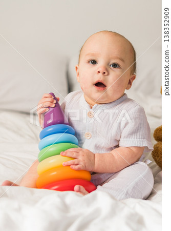 Cute baby playing with colorful rainbow toy pyramid and teddy bear sitting on bed in white sunny bedroom copy space. Toys for little kids. Child with educational toy. Cute baby playing with colorful rainbow toy pyramid and teddy bear sitting on bed in white sunny bedroom copy space. Toys for little kids. Child with educational toy. 138352909