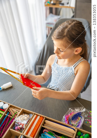 Little girl admiring finished handmade craft made from wooden sticks and colorful materials at table. Kids creativity imagination development and learning through play concept. Little girl admiring finished handmade craft made from wooden sticks and colorful materials at table. Kids creativity imagination development and learning through play concept. 138353055