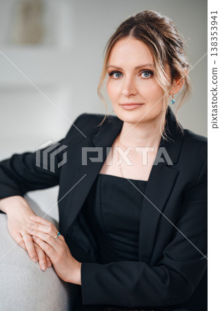 Woman in black suit sits on a couch with a soft expression indoors Woman in black suit sits on a couch with a soft expression indoors 138353941