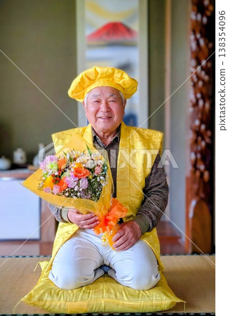 一位身穿黃色喜慶服裝、手捧鮮花、面帶微笑的老者，在日式房間裡慶祝他的八十歲生日。 （紀念照） 138354096