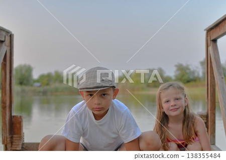 A little boy and cute girl are sitting on a wooden pier on the bank of a lake. Small children are sitting on a pier at the side of a pond. The concept of a carefree childhood and summer holidays. A little boy and cute girl are sitting on a wooden pier on the bank of a lake. Small children are sitting on a pier at the side of a pond. The concept of a carefree childhood and summer holidays. 138355484