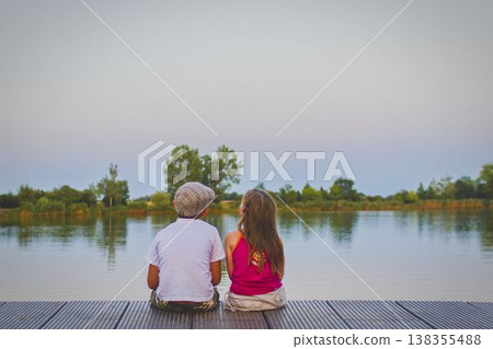 A little boy wearing a flat cap and a cute little girl sit with their backs turned on a wooden pier on the shore of a lake. Small children sit on a pier at the edge of a pond. The concept of a A little boy wearing a flat cap and a cute little girl sit with their backs turned on a wooden pier on the shore of a lake. Small children sit on a pier at the edge of a pond. The concept of a 138355488