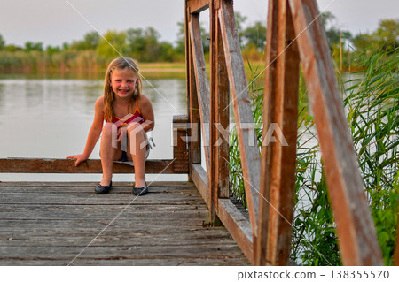 Young girl in swimsuit and shoes sitting on a wooden pier by the lake, smiling Young girl in swimsuit and shoes sitting on a wooden pier by the lake, smiling 138355570