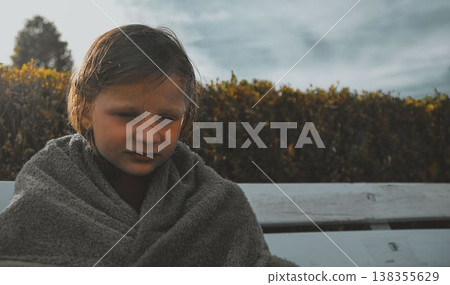 A sad little girl covered in a towel sits on a bench at the swimming pool. The concept of summer vacations, freedom and childhood. A sad little girl covered in a towel sits on a bench at the swimming pool. The concept of summer vacations, freedom and childhood. 138355629