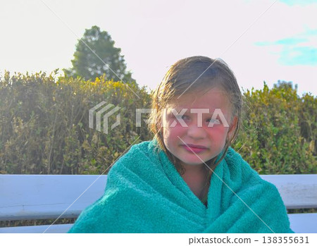 A little laughing girl wearing a towel is sitting on a bench at the pool. The concept of summer vacation, carefree days, and a happy childhood. A little laughing girl wearing a towel is sitting on a bench at the pool. The concept of summer vacation, carefree days, and a happy childhood. 138355631