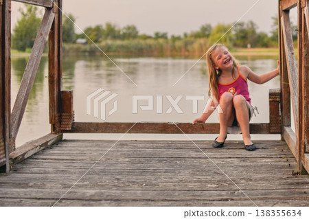 A little laughing girl sits on a wooden pier at the shore of a lake. The photo was taken during the golden hour, at sunset. The concept of childhood, summer holidays and carefree days. A little laughing girl sits on a wooden pier at the shore of a lake. The photo was taken during the golden hour, at sunset. The concept of childhood, summer holidays and carefree days. 138355634