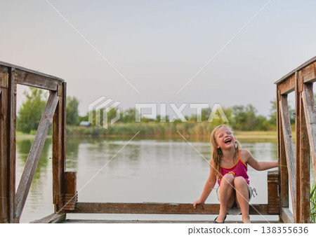 Joyful young girl laughing heartily while sitting on a wooden pier by the lake. A happy child with eyes closed in laughter sits on a rustic wooden dock, surrounded by serene natural scenery and calm Joyful young girl laughing heartily while sitting on a wooden pier by the lake. A happy child with eyes closed in laughter sits on a rustic wooden dock, surrounded by serene natural scenery and calm 138355636