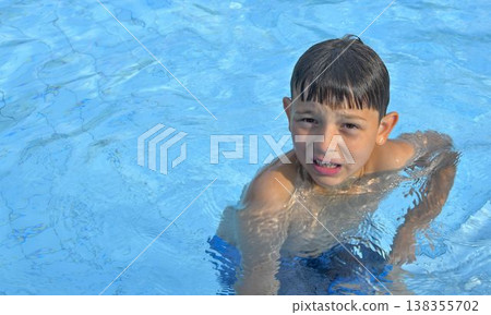 A cute little boy at the swimming pool. A smiling little kid in the water at the swimming pool. The concept of summer, summer holidays, rest, and relaxation. A cute little boy at the swimming pool. A smiling little kid in the water at the swimming pool. The concept of summer, summer holidays, rest, and relaxation. 138355702