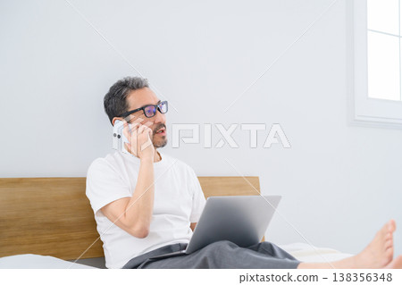 A Japanese man talking on the phone while using his laptop in bed. A Japanese man talking on the phone while using his laptop in bed. 138356348