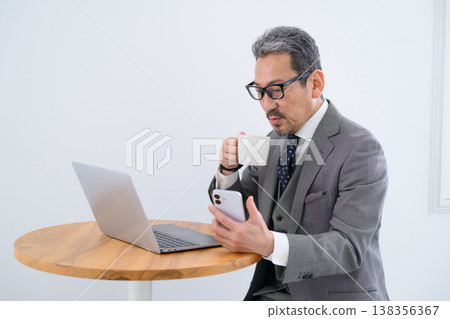 A Japanese man is working on his laptop while looking at his smartphone at a table. A Japanese man is working on his laptop while looking at his smartphone at a table. 138356367