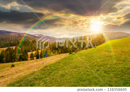 sunset over mountain scenery with coniferous forest. countryside landscape with grassy fields and rolling hills. springtime rural area under cloudy sky and rainbow. wonderful outdoor adventure 138356695