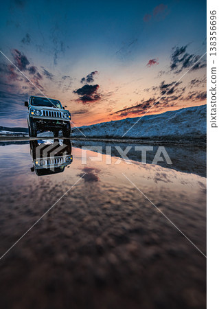 A four-wheel drive vehicle stands on a snowy road, its reflection against the twilight sky. 138356696