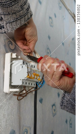 A man's hands hold pliers and secure the wires going to a wall-mounted home electrical distribution box with circuit breakers. DIY wall-mounted electrical equipment installation using a hand tool. 138356802