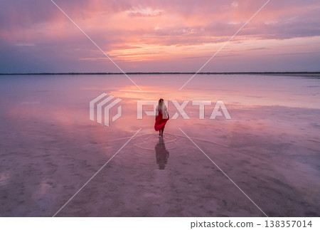 Aerial of a young woman in red dress walking in the water of a unique pink salt lake. Sunset at lake Bursol with beautiful reflections on calm water surface. Stunning scenery Aerial of a young woman in red dress walking in the water of a unique pink salt lake. Sunset at lake Bursol with beautiful reflections on calm water surface. Stunning scenery 138357014