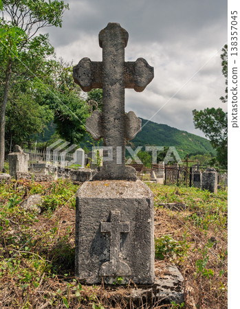 Ancient stone cross monument rises above overgrown grass in a quiet rural cemetery on a cloudy afternoon Ancient stone cross monument rises above overgrown grass in a quiet rural cemetery on a cloudy afternoon 138357045