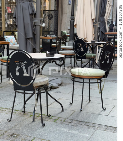 Empty street cafe terrace with wrought iron chairs and closed umbrellas near shopfronts. Outdoor hospitality layout, pedestrian dining zone, urban leisure setting, and calm city center atmosphere Empty street cafe terrace with wrought iron chairs and closed umbrellas near shopfronts. Outdoor hospitality layout, pedestrian dining zone, urban leisure setting, and calm city center atmosphere 138357208