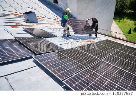 Workers building solar panel system on metal rooftop of house. Two men installers carrying photovoltaic solar module outdoors. Alternative, green and renewable energy generation concept. 138357687