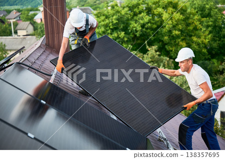 Workers building solar panel system on roof of house. Two men installers in helmets carrying photovoltaic solar module outdoors. Alternative, green and renewable energy generation concept. Workers building solar panel system on roof of house. Two men installers in helmets carrying photovoltaic solar module outdoors. Alternative, green and renewable energy generation concept. 138357695
