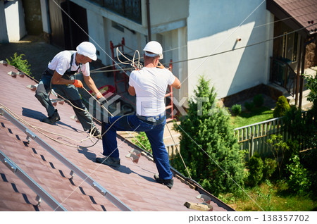 Men workers installing solar panel system on roof of house. Electricians in helmets lifting up photovoltaic solar module with help of ropes outdoors. Concept of alternative and renewable energy. 138357702