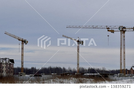 Three enormous construction cranes are actively operating in a winter landscape beneath an overcast, cloudy sky 138359271