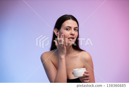 Young woman applying face cream, moisturizing and caring for her skin, promoting beauty, wellness, and self-care routines for healthy complexion against a vibrant purple and pink background Young woman applying face cream, moisturizing and caring for her skin, promoting beauty, wellness, and self-care routines for healthy complexion against a vibrant purple and pink background 138360885