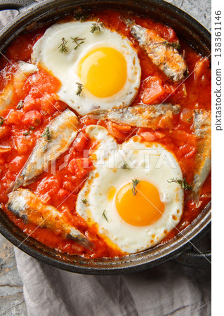 Light breakfast fried eggs with sardines in spicy tomato sauce close-up in a frying pan on the table. Vertical top view Light breakfast fried eggs with sardines in spicy tomato sauce close-up in a frying pan on the table. Vertical top view 138361146