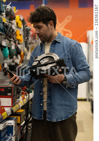 Man holding circular saw and smartphone, researching tools in hardware store 138361167